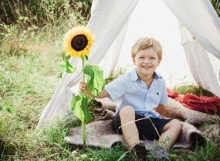 Ein kleiner Junge sitzt lächelnd unter einem Kinderzelt im Garten und hält eine Sonnenblume.
