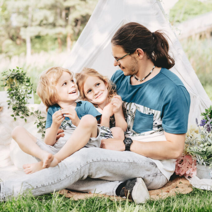 Ein Vater mit seinen beiden Kindern auf einer hellen Decke im Grünen, vor einem hellen Tipi