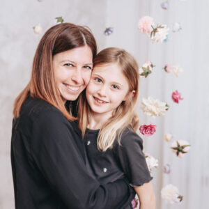 Foto einer Mama und ihrer Tochter, aneinander gekuschelt in die Kamera lachend, umgebene von vielen schwebenden Blüten