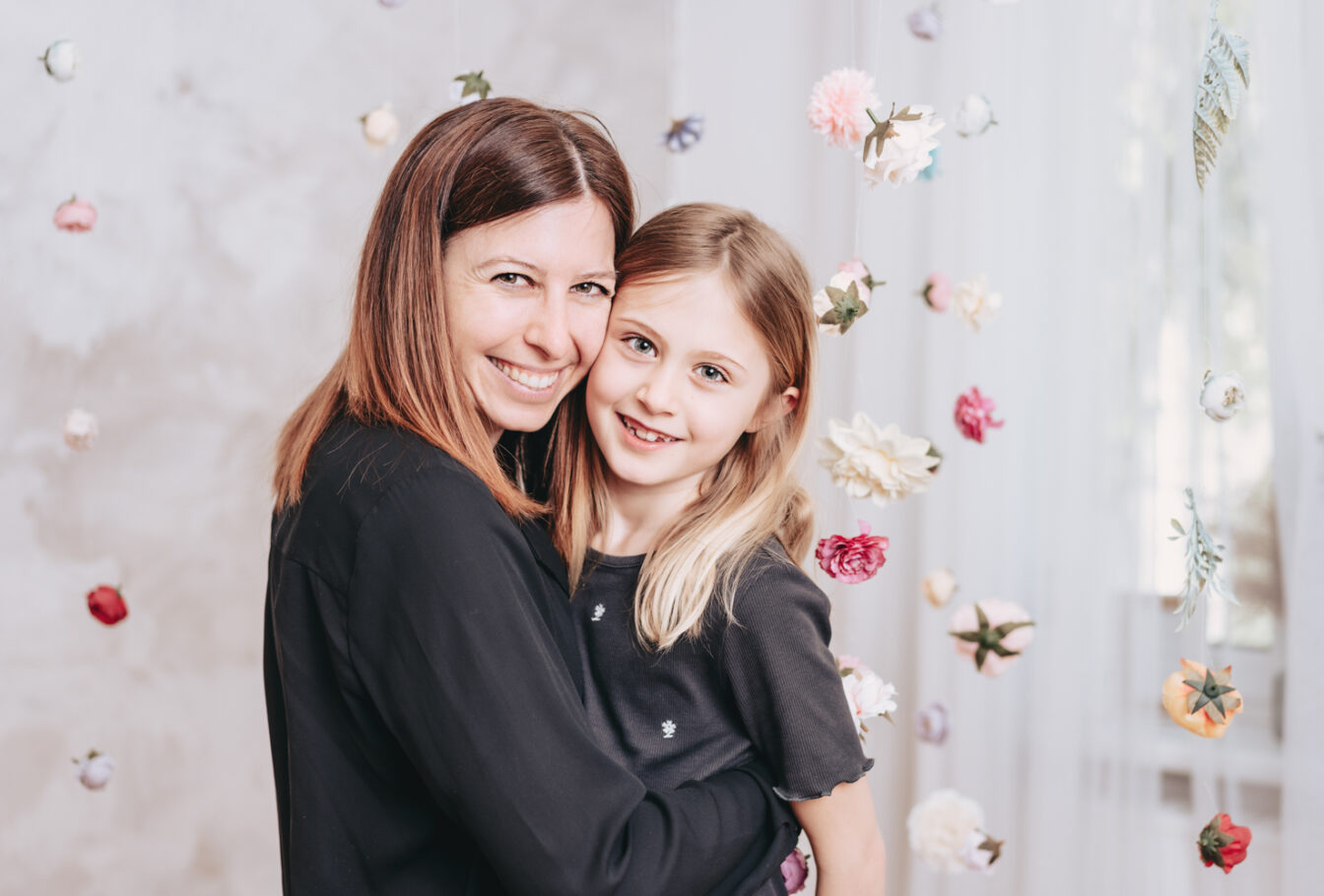 Foto einer Mama und ihrer Tochter, aneinander gekuschelt in die Kamera lachend, umgebene von vielen schwebenden Blüten