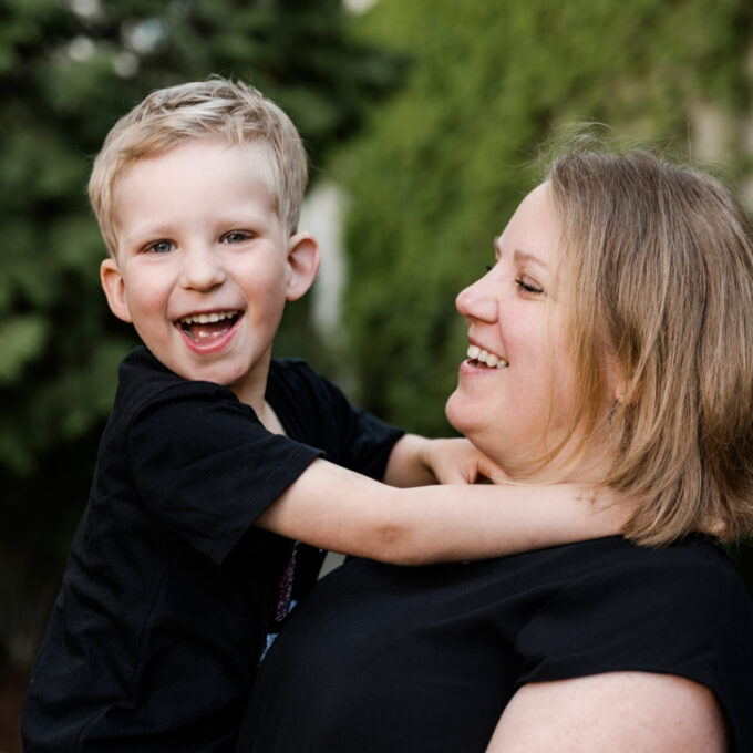 Foto einer Mama im Grünen, mit ihrem Sohn auf dem Arm, wobei das Kind in die Kamera lacht