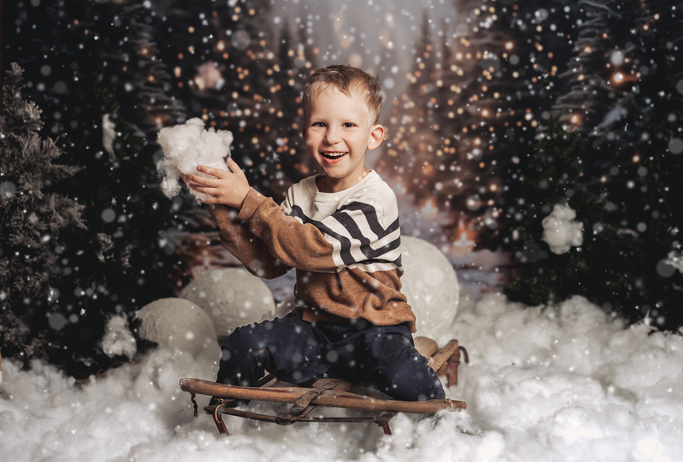 Weihnachtsfoto eines Jungen auf einem Schlitten, in einem abendlichen Wald, der mit Schnee spielt