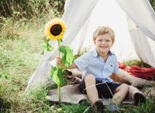 Ein kleiner Junge sitzt l&auml;chelnd unter einem Kinderzelt im Garten und h&auml;lt eine Sonnenblume.
