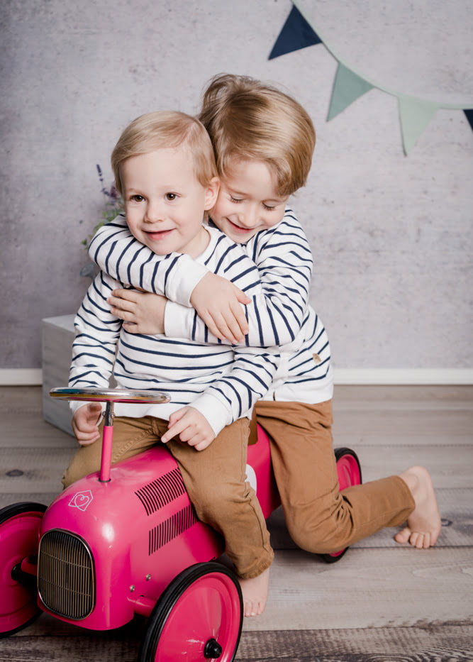 Ein &auml;lteres Gewschisterkind umarmt das j&uuml;ngere, welches auf einem pinken Traktor sitzt.
